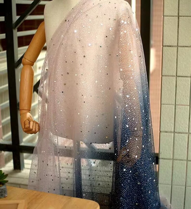 K&ouml;nnte beinhalten: Ein transparenter Stoff mit Farbverlauf, der von einem zarten Rosa zu einem tiefen Marineblau &uuml;bergeht. Der Stoff ist mit silbernen Sternen und Mondformen verziert, wodurch ein himmlisches Muster entsteht. Der Stoff ist &uuml;ber einer Schaufensterpuppe drapiert.