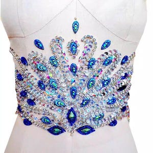 May include: A white fabric with a large blue and clear crystal embellishment in the shape of a peacock feather. The embellishment is made of rhinestones and sequins.