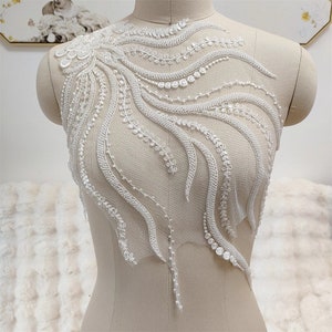 以下が含まれることがあります： 繊細な花のデザインが施された白いビーズレースのアップリケ。アップリケはビーズとスパンコールの渦巻き模様で、豪華でエレガントな外観を作り出しています。