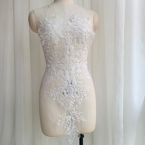 May include: A white floral lace applique featuring intricate embroidery of leaves, vines, and blossoms. The design is symmetrical, extending vertically down the center of a sheer, translucent mesh fabric base, creating a detailed botanical pattern.