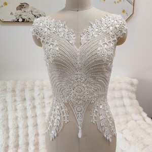 May include: White beaded and lace applique with a V-neckline and a pointed bottom. The applique is on a dress form.