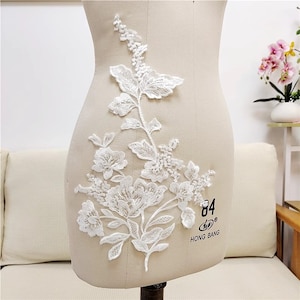 Könnte beinhalten: Weiße Spitzenapplikation mit floralem Design. Die Applikation zeigt eine große Blume mit kleineren Blumen und Blättern. Die Applikation befindet sich auf weißem Hintergrund. Der Text "84 HONG BANG" ist in der unteren rechten Ecke sichtbar.