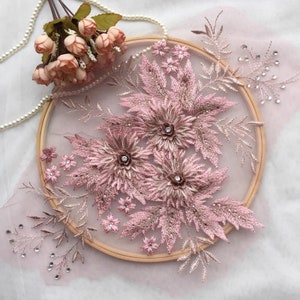May include: A pink floral applique with rhinestones and glitter on a sheer fabric background. The applique is displayed on a wooden embroidery hoop.