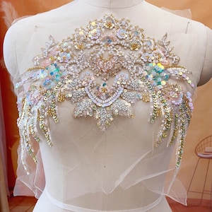 May include: A delicate white and gold beaded applique with iridescent sequins and pearls. The applique features a floral design with a central heart shape.