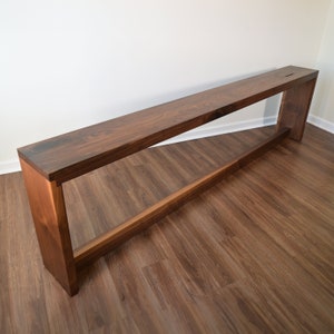 May include: A long, narrow wooden console table with a dark brown finish. The table has a simple, modern design with a single shelf and a rectangular top.