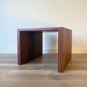 May include: A dark brown, rectangular wooden side table with a U-shaped design. The table is made of solid wood and has a smooth, polished finish. The table is approximately 18 inches long, 12 inches wide, and 16 inches tall.