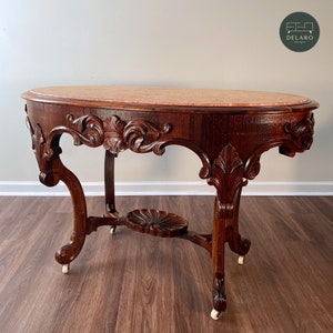 May include: A vintage oval-shaped table with a carved wooden base and a pink marble top. The table has four legs with intricate details and wheels.