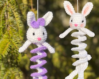 May include: Two crocheted bunny ornaments hanging from a tree branch. One bunny is white with a purple spiral body and a purple bow. The other bunny is white with a white spiral body. The text "Crafty Crochet Castle" is visible at the bottom of the image.