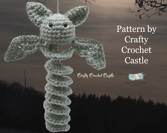 Puede incluir: Un móvil de viento de murciélago de crochet gris colgado de una rama de árbol. El murciélago tiene un cuerpo en espiral y está diseñado para girar con el viento. El texto "Pattern by Crafty Crochet Castle" es visible en la imagen.