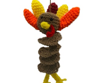 May include: A crocheted turkey ornament with a brown body, yellow legs, and a red wattle. The turkey has a yellow, orange, and red feathered head.