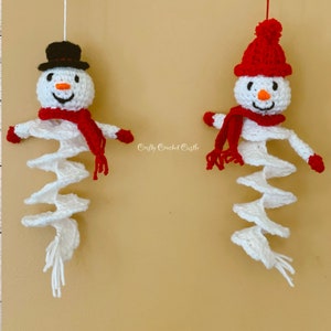 May include: Two crocheted snowman ornaments hanging from white string. The snowman on the left has a black top hat and a red scarf. The snowman on the right has a red knit hat with a white pom pom and a red scarf. Both snowmen have white crocheted bodies.