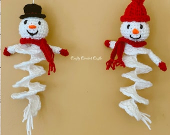 May include: Two crocheted snowman ornaments hanging from white string. The snowman on the left has a black top hat and a red scarf. The snowman on the right has a red knit hat with a white pom pom and a red scarf. Both snowmen have white crocheted bodies.