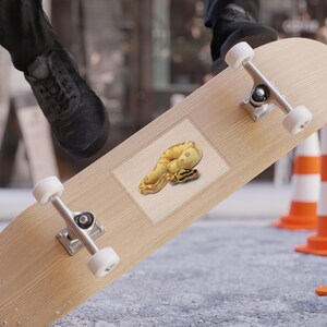 Può includere: Uno skateboard marrone chiaro con hardware argentato e ruote bianche. Un adesivo con un disegno di serpente giallo e marrone è sulla tavola. Lo sfondo include coni stradali e le gambe e i piedi di una persona.