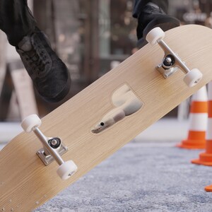 Può includere: Uno skateboard marrone chiaro con carrelli argentati e ruote bianche. Un adesivo di un serpente bianco è sul ponte. Lo skateboard è angolato, con la scarpa nera di una persona visibile sullo sfondo, insieme a coni stradali arancioni e bianchi.