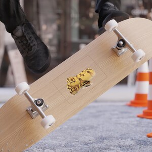 Può includere: Un adesivo con un disegno di serpente giallo e marrone è posto su uno skateboard. Lo skateboard è marrone chiaro con hardware argentato e ruote bianche. Lo sfondo include una scarpa nera e coni arancioni.