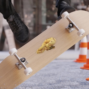 May include: A yellow and black sticker of a snake on a skateboard. The sticker is a detailed illustration of a snake with a coiled body. The skateboard is made of light-colored wood and has silver trucks and white wheels. The background includes construction cones.