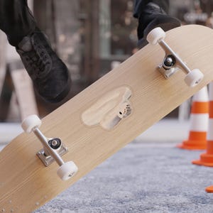 Può includere: Uno skateboard in legno di colore chiaro con ruote bianche e carrelli argentati. La tavola ha un design ritagliato con una piccola figura di colore chiaro all'interno. Lo skateboard è su una superficie grigia con coni stradali arancioni e bianchi sullo sfondo.