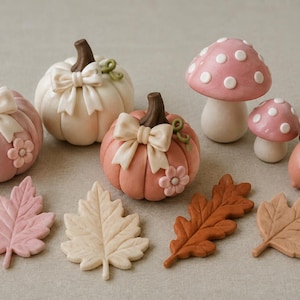 May include: A collection of decorative clay figures including two pink pumpkins and one cream pumpkin, each with a bow and flower detail. Two pink and white spotted mushrooms, one brown acorn with a flower, and four leaves in pink, cream, and brown.