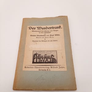 Könnte beinhalten: Ein alter Buchumschlag mit dem Titel "Der Wundertrank" in schwarzer Schrift. Der Umschlag ist hellbraun und hat einen verblassten, abgenutzten Look. Das Buch ist ein Schauspiel mit Musik von Walter Stratmann und Paul Wille.