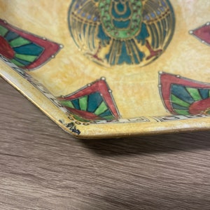 May include: A yellow ceramic bowl with a multi-coloured design featuring a stylised bird with green, blue, and gold accents. The bowl has a geometric pattern around the rim.