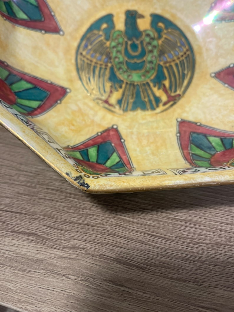 May include: A yellow ceramic bowl with a multi-coloured design featuring a stylised bird with green, blue, and gold accents. The bowl has a geometric pattern around the rim.