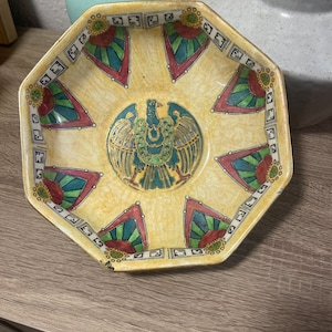 May include: Octagonal ceramic bowl with a yellow background and a colourful geometric design. The centre of the bowl features a green, gold, and black stylised bird.