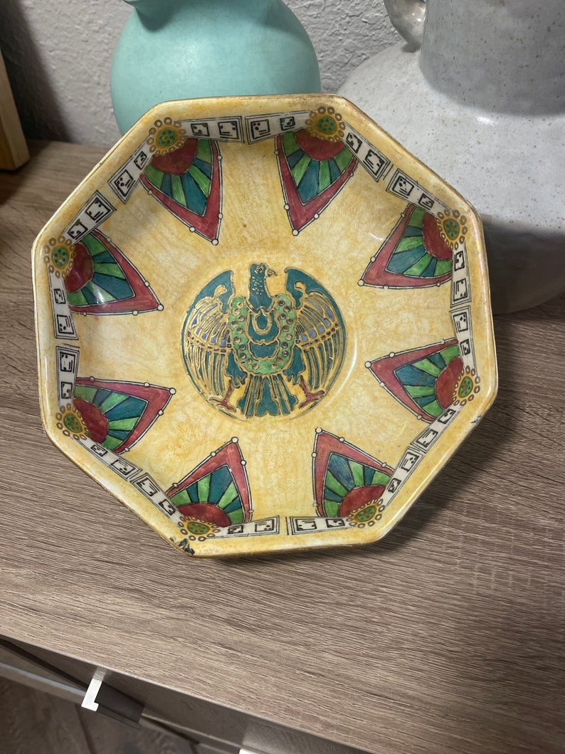 May include: Octagonal ceramic bowl with a yellow background and a colourful geometric design. The centre of the bowl features a green, gold, and black stylised bird.
