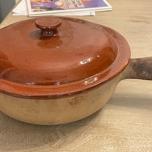 Puede incluir: Una olla de cerámica marrón con tapa y asa de madera. La olla tiene un esmalte marrón claro en la parte inferior y un esmalte marrón oscuro en la parte superior. La tapa tiene un pequeño pomo en la parte superior.