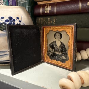 May include: An antique framed photograph of a person in a dark dress with a patterned shawl. The photograph is in a gold-colored frame and encased in a black, hinged case. The image is displayed on a light blue shelf with books and a beaded necklace.