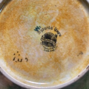 May include: Close-up of the underside of a ceramic plate with a brown glaze. The plate features a black and white logo that reads "W. Bizanta Ware" and "Pottery Made in England". The number "113" is written in black on the base of the plate.