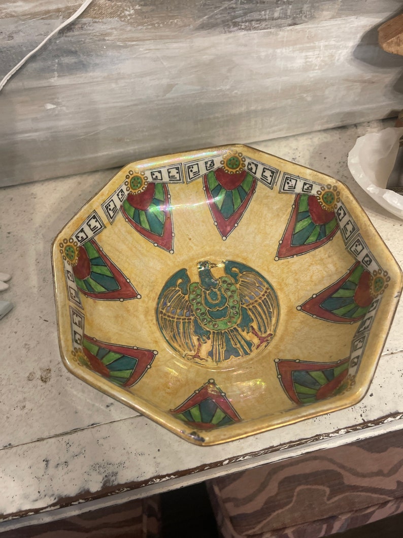 May include: Octagonal ceramic bowl with a yellow glaze and a colourful design. The design features a stylised bird with green, red, and blue accents. The bowl has a gold rim.