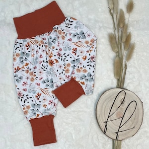 Könnte beinhalten: Weiße Hose mit Blumenmuster in Orange-, Rosa- und Grautönen. Die Hose hat einen rostfarbenen Bund und Bündchen.