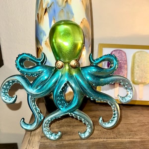 May include: A decorative octopus figurine with a metallic teal and green finish. The octopus has eight curling tentacles and a rounded head. The figurine has gold-colored accents and is displayed on a wooden surface.