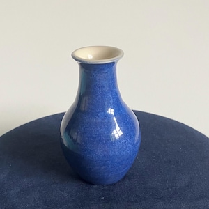 May include: A small, blue ceramic vase with a speckled texture. The vase has a rounded body and a narrow neck, with a cream-coloured interior. The vase is sitting on a dark blue surface. A decorative item for the home.