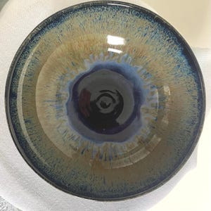 May include: A handcrafted ceramic bowl with a glossy finish. The bowl has a dark blue centre that fades into lighter blue and tan shades towards the rim. The rim is a darker blue. The bowl is round and has a smooth interior.
