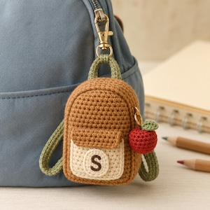 Puede incluir: Un llavero de mochila tejido a ganchillo en color marrón con asa y correas verde claro, que presenta un bolsillo delantero color crema con una letra S marrón oscuro y un pequeño dije de manzana roja tejido a un lado con un broche dorado.