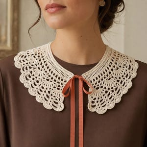 May include: A cream-colored crocheted detachable collar featuring an intricate scalloped lace pattern and a delicate edge. The collar is secured at the center front with a thin, dusty rose-colored ribbon tied into a simple bow, resting on a brown shirt.