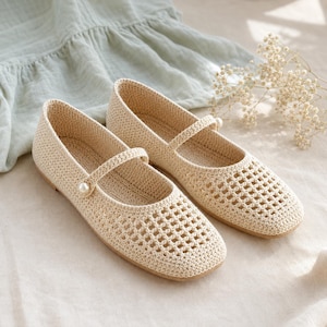 May include: A pair of beige crochet flats featuring a square toe, a delicate strap across the instep, and a single white pearl accent on each side. The shoes have a woven texture with a grid-like pattern on the front and a solid heel area.