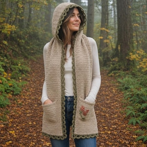 May include: A beige crochet hooded scarf with a dark olive green scalloped trim along the edges. The garment features two front pockets, with the right pocket displaying a small, embroidered floral design in shades of orange and green.