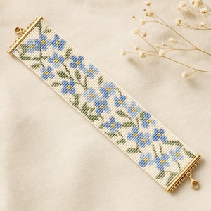 May include: A wide beaded bracelet featuring a repeating floral pattern of light blue petals with yellow centers and green stems on an off-white background. The bracelet is finished with gold-toned metal end bars and a lobster claw clasp.