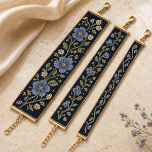 May include: Three beaded bracelets of varying widths featuring a dark navy background with light blue floral patterns, green leaves, and gold-toned metal end bars with lobster claw clasps and adjustable chain links for a secure fit.
