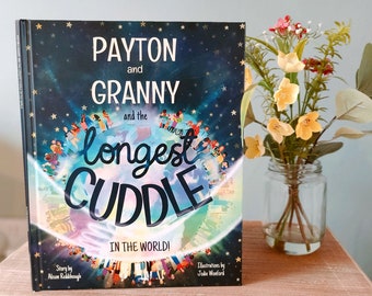 May include: A children's book titled "Payton and Granny and the Longest Cuddle in the World!" features a starry night sky background with a globe illustration. The book is displayed on a wooden surface next to a glass jar with flowers.