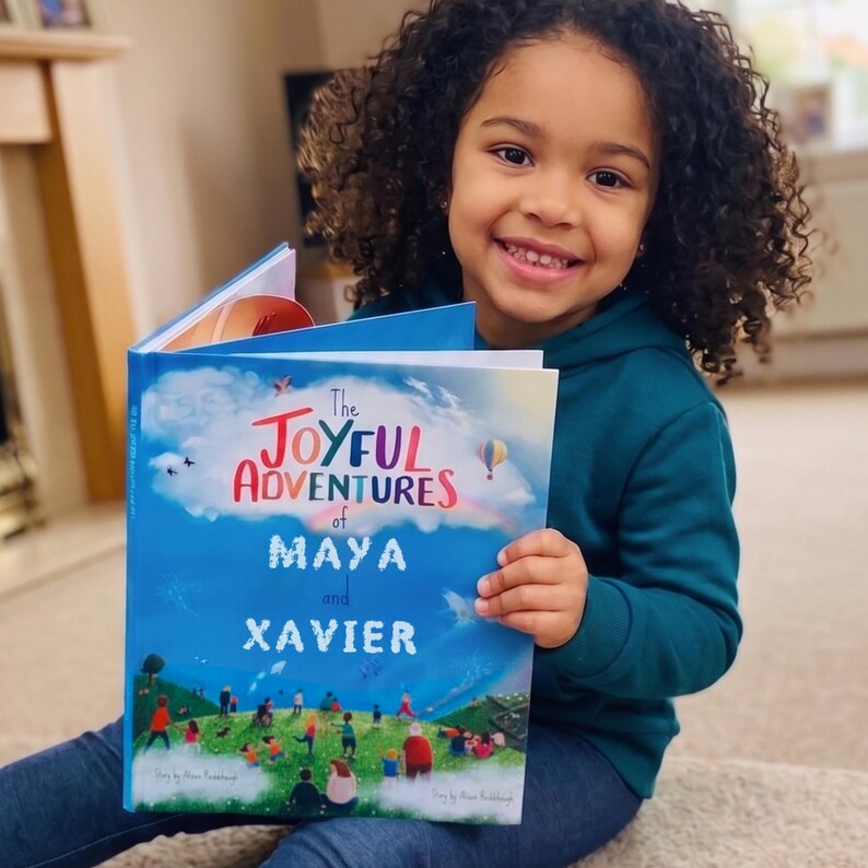 May include: A young child smiles while holding a children's book titled "The Joyful Adventures of Maya and Xavier." The book cover features a blue sky with colorful illustrations and text. The child is wearing a teal hoodie.