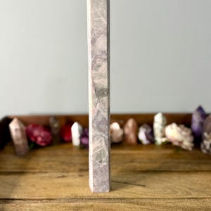 May include: A tall, light purple crystal obelisk with a pointed top. The obelisk has a marbled appearance with streaks of white and green. It stands on a wooden surface, with other crystals and flowers in the background.