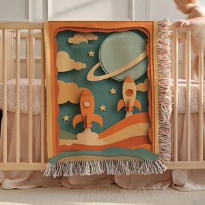 May include: A woven baby blanket with a space-themed design. The blanket features two orange rockets launching, a planet with a ring, clouds, and stars. The blanket has a fringed edge and is displayed on a wooden crib.