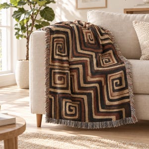 May include: A woven throw blanket featuring a repeating geometric pattern of interlocking brown, black, and tan squares and zigzags. The rectangular textile has a textured surface and is finished with a short, frayed fringe along the edges.