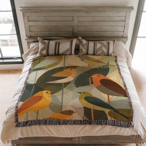 May include: A woven throw blanket featuring a colorful bird and leaf design. The blanket showcases birds in shades of yellow, orange, and brown against a backdrop of green and tan leaves. The blanket has a fringed edge and is displayed on a bed.