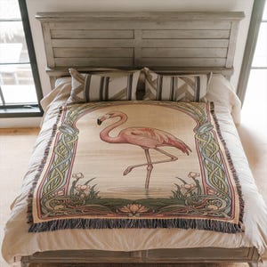Puede incluir: Una manta decorativa con un flamenco rosa sobre un fondo crema, enmarcada por un arco con diseños florales y de nudos celtas. La manta, con bordes con flecos, está colocada sobre una cama con almohadas a rayas.