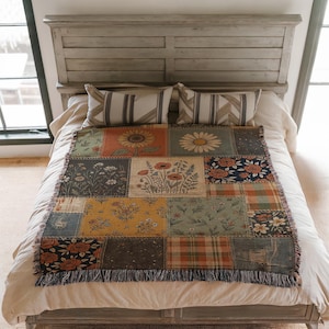 Op de afbeelding: Een patchwork sprei met bloemen en patronen. De sprei heeft zonnebloemen, madeliefjes en andere botanische ontwerpen in oranje, geel, groen en blauw. De sprei heeft een gefranjerde rand en wordt op een bed getoond.