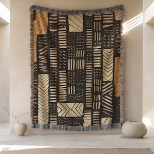 May include: A rectangular woven throw blanket featuring a dark charcoal background with a repeating pattern of tan and cream-colored geometric shapes, including vertical stripes, chevrons, and dashes, finished with a fringed edge on all sides.
